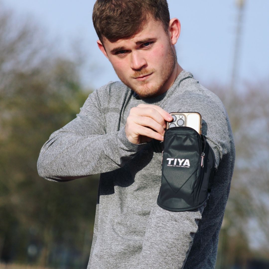 A man in a gray athletic shirt securing a “TIYA” running arm phone holder to his arm while standing outside.